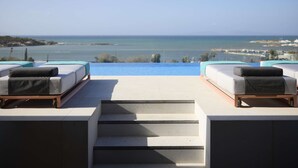 A rooftop terrace with two lounge chairs, a swimming pool, and a view of the sea and distant buildings. - Dusit Suites Athens (Glyfada)