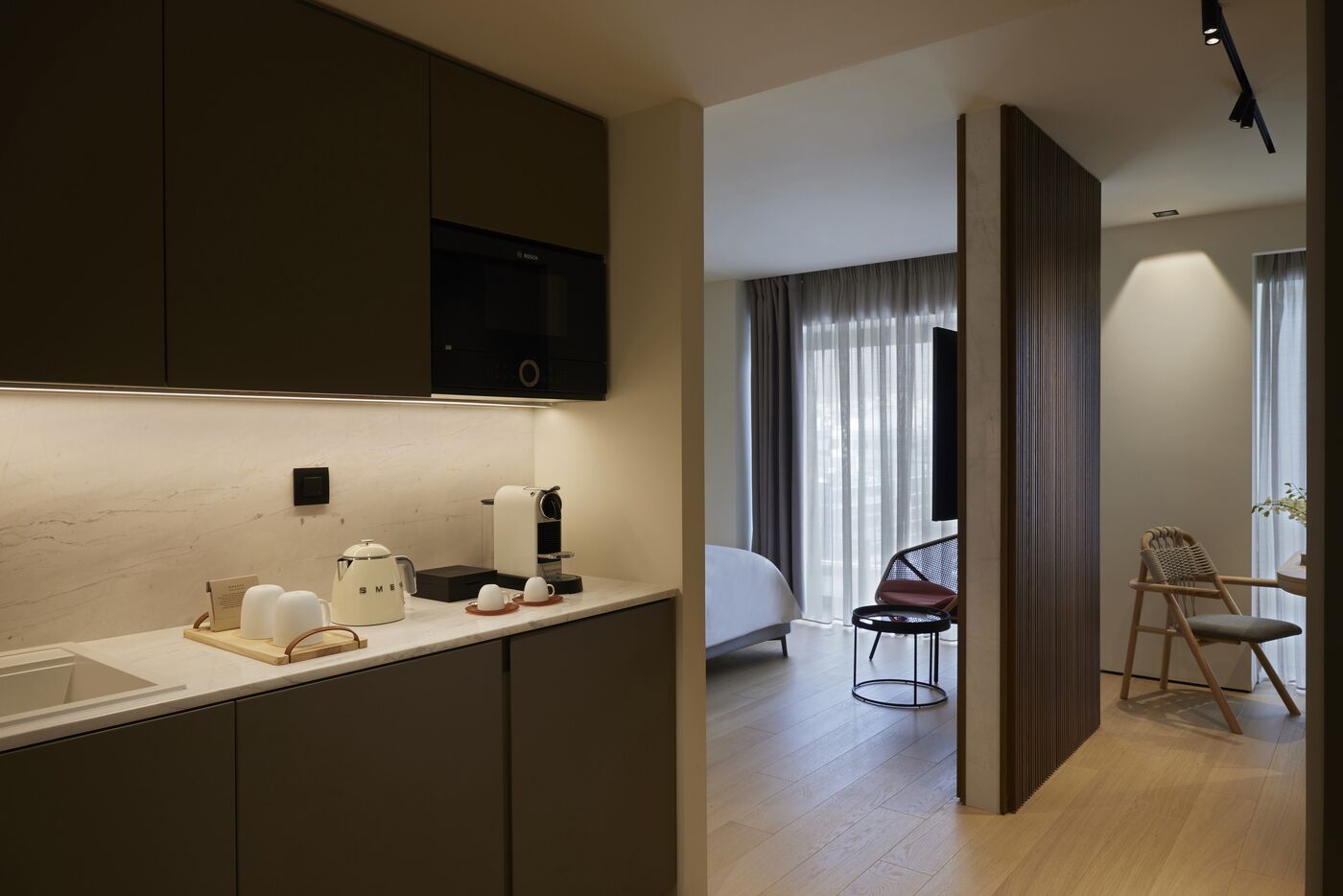Una habitación de hotel moderna con kitchenette, una cama, zona de estar con una silla y mesa, y un ventanal con cortinas.