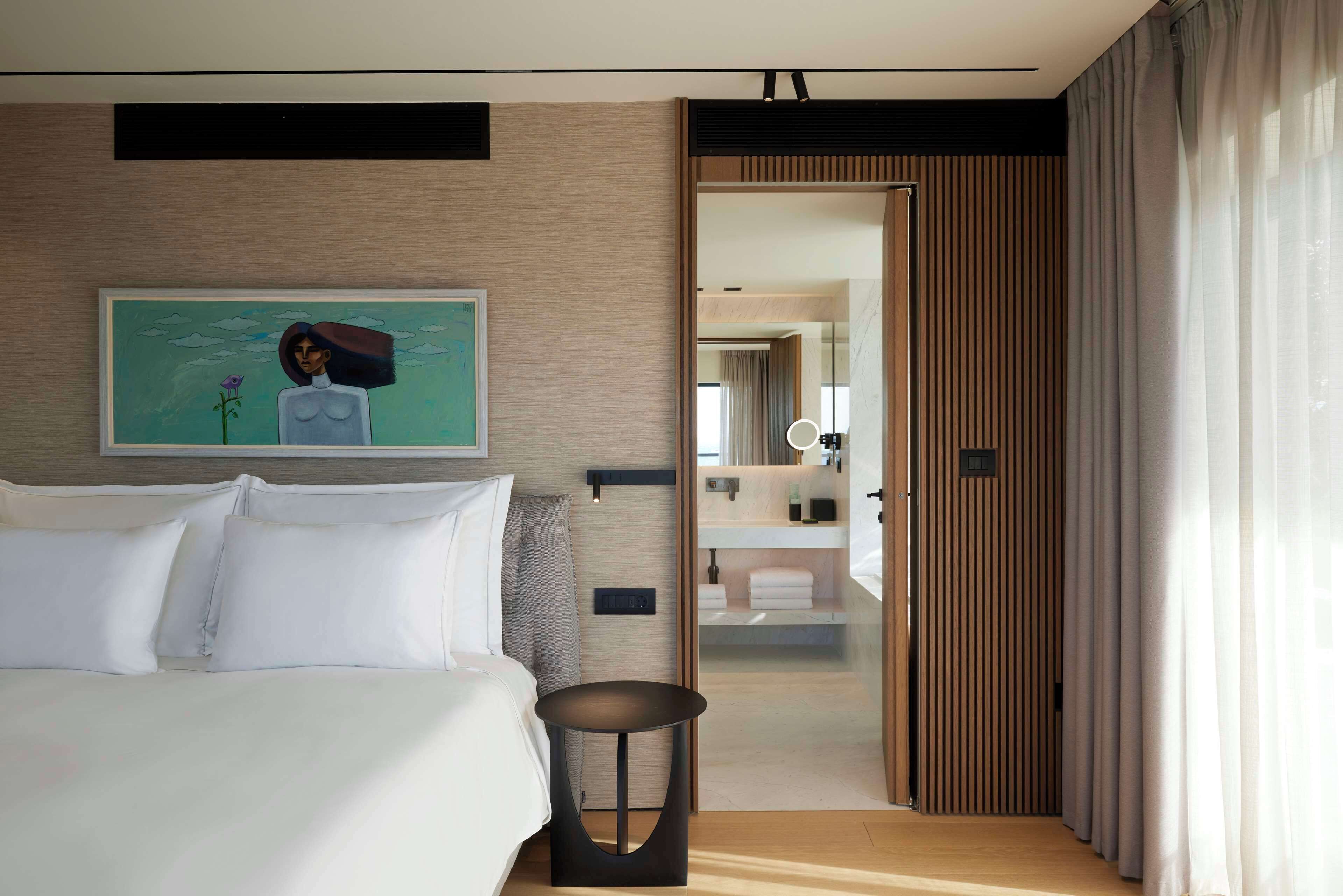 Une chambre d’hôtel moderne avec un grand lit, une petite table de chevet, un tableau au mur et une vue sur la salle de bain.