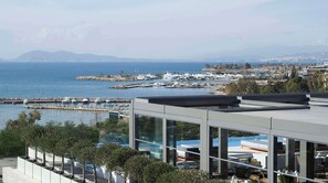A modern building with a glass facade overlooking a marina with boats. - Dusit Suites Athens (Glyfada)