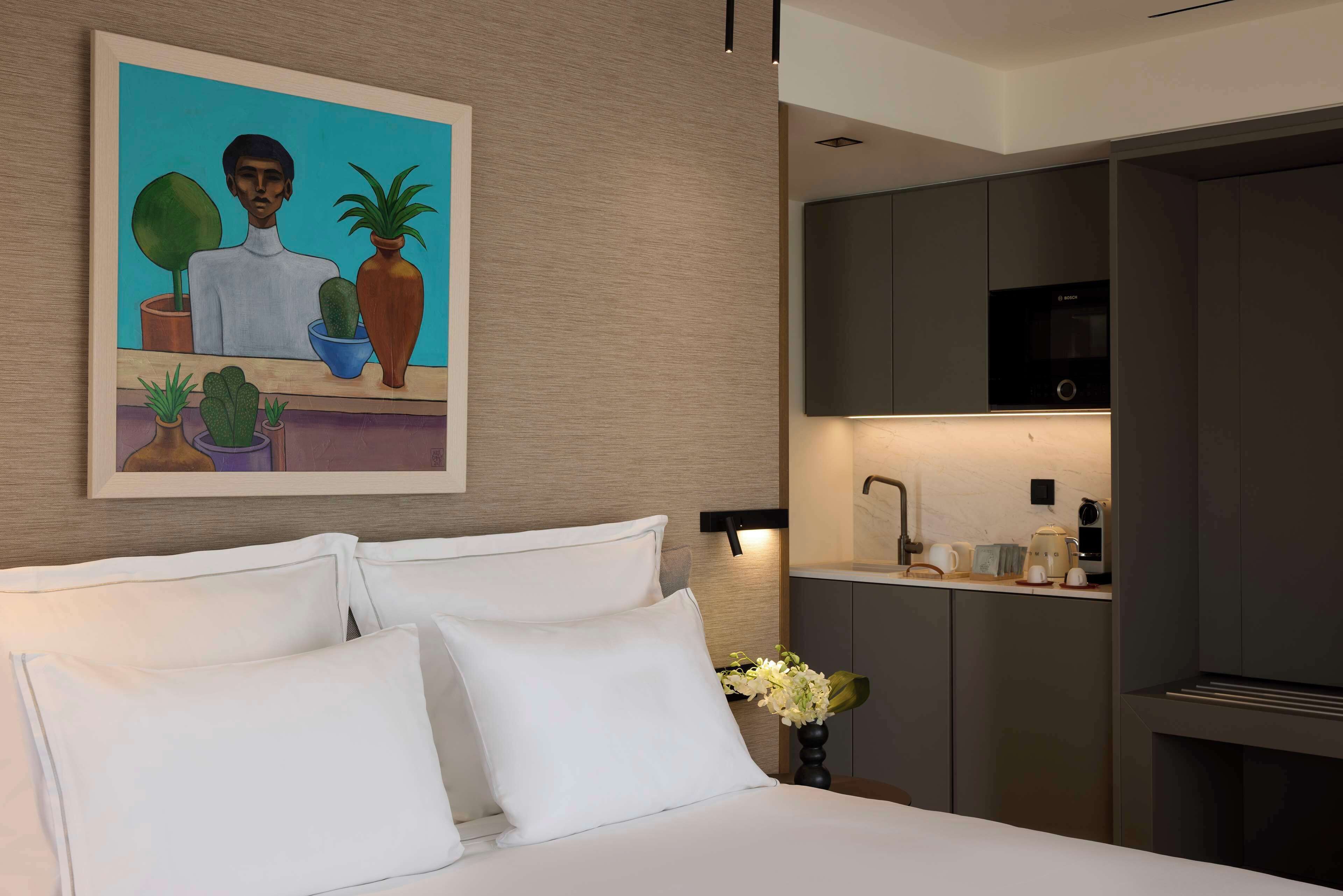 Una habitación de hotel moderna con una cama, un cuadro, una kitchenette y una mesita de noche con un jarrón de flores.