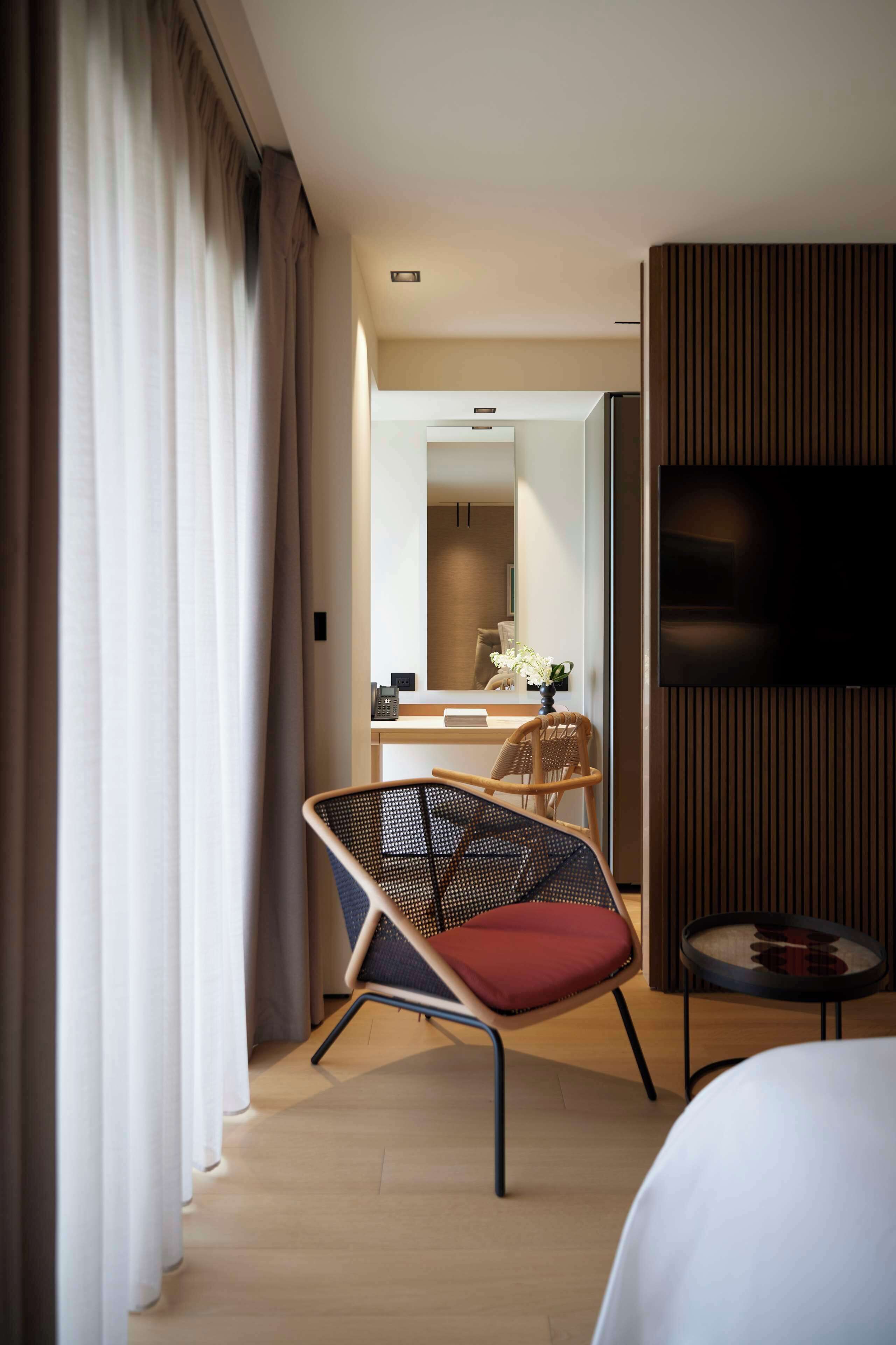 Ein Hotelzimmer mit Bett, Sessel mit rotem Kissen, einem kleinen runden Tisch, einem an der Wand befestigten Fernseher und einem Spiegel, der den Raum widerspiegelt.
