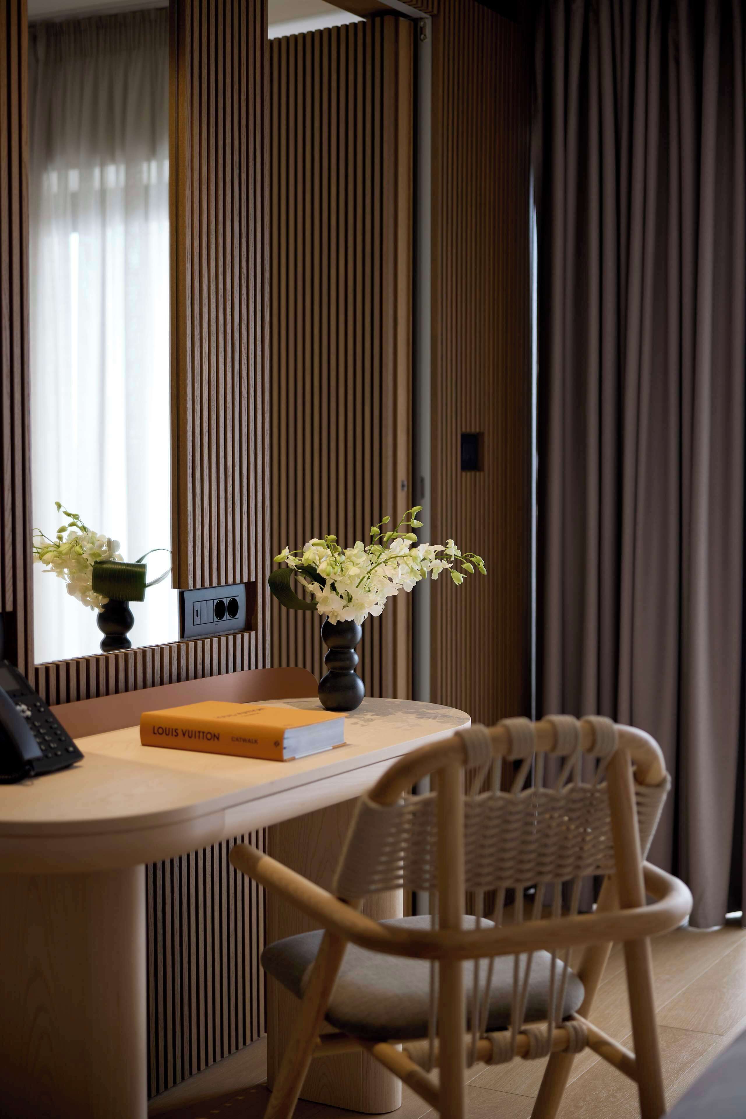 Ein Hotelzimmer mit einem Holztisch, einem Stuhl, einer Vase mit Blumen, einem Buch mit dem Titel 'LOUIS VUITTON' und einem Spiegel.