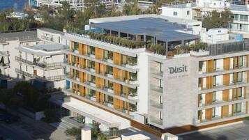 A modern multi-story hotel with a prominent 'Dusit Suites - Athens' sign, featuring balconies with plants, a rooftop terrace, and a glass facade.