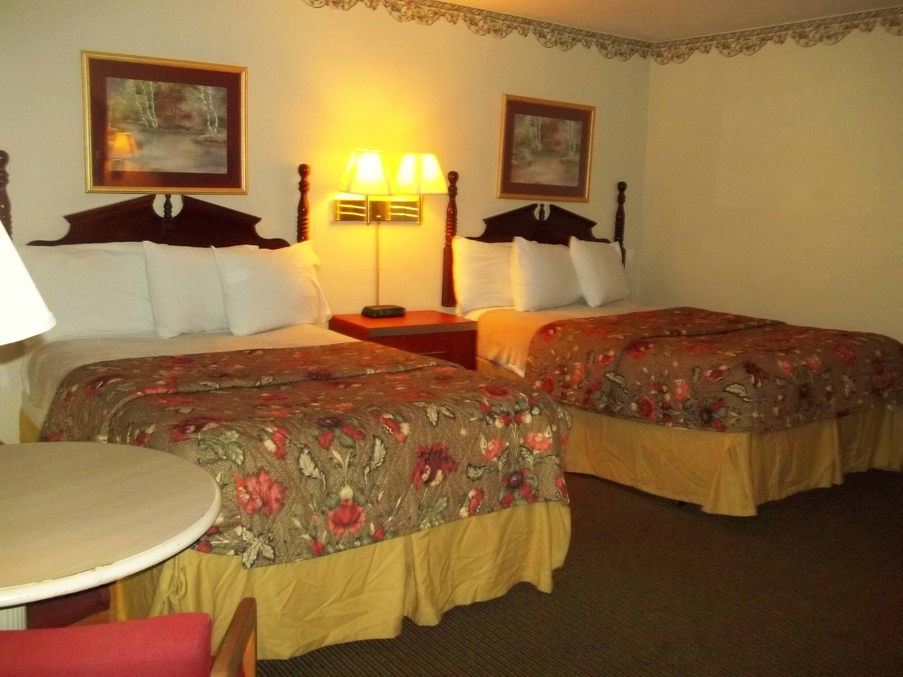 A hotel room with two beds, a small table, and a chair. There are two lamps on wooden nightstands, and the walls are decorated with framed pictures.
