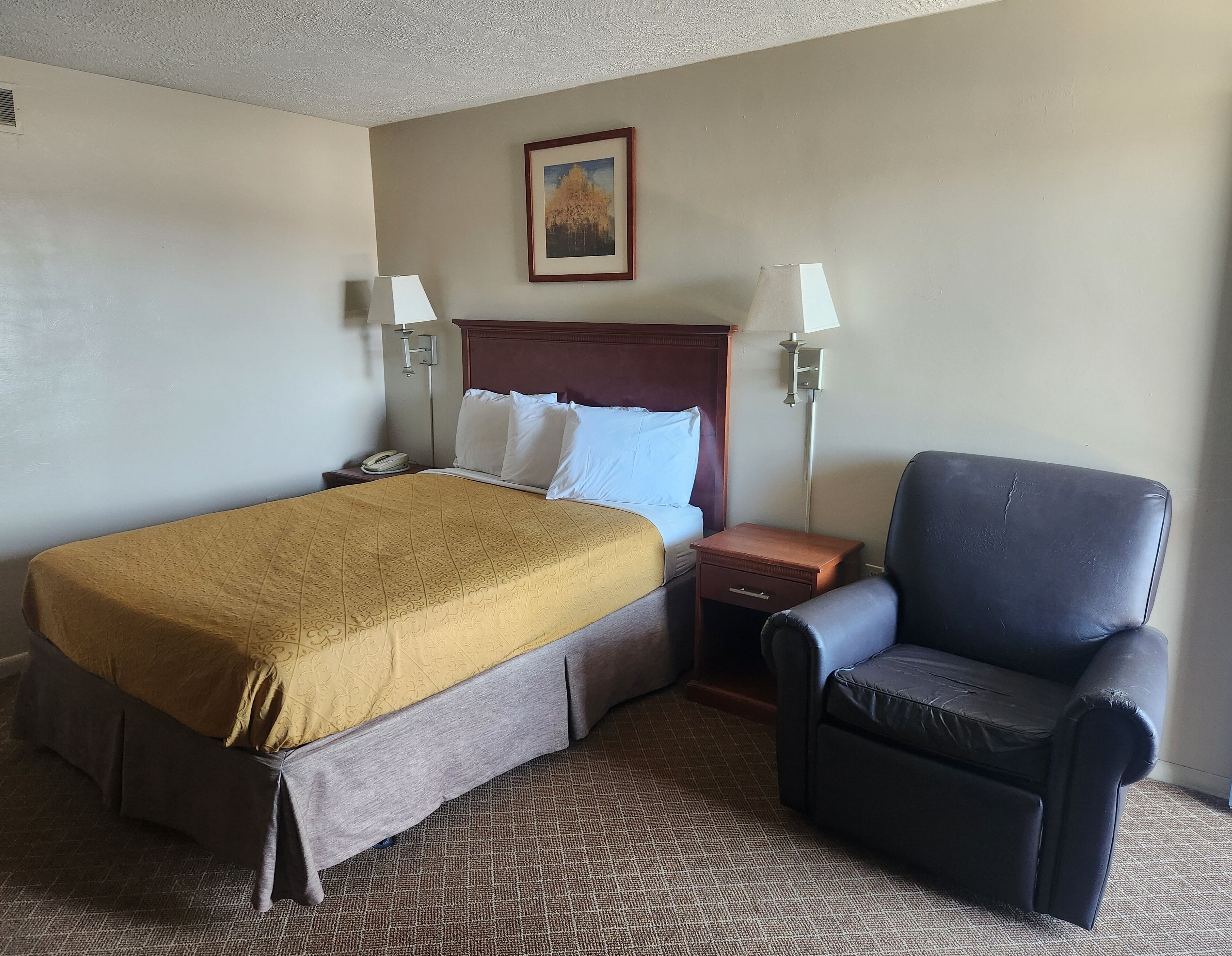 Habitación de hotel con una cama grande, dos mesitas de noche, una silla, un cuadro en la pared y suelo alfombrado.