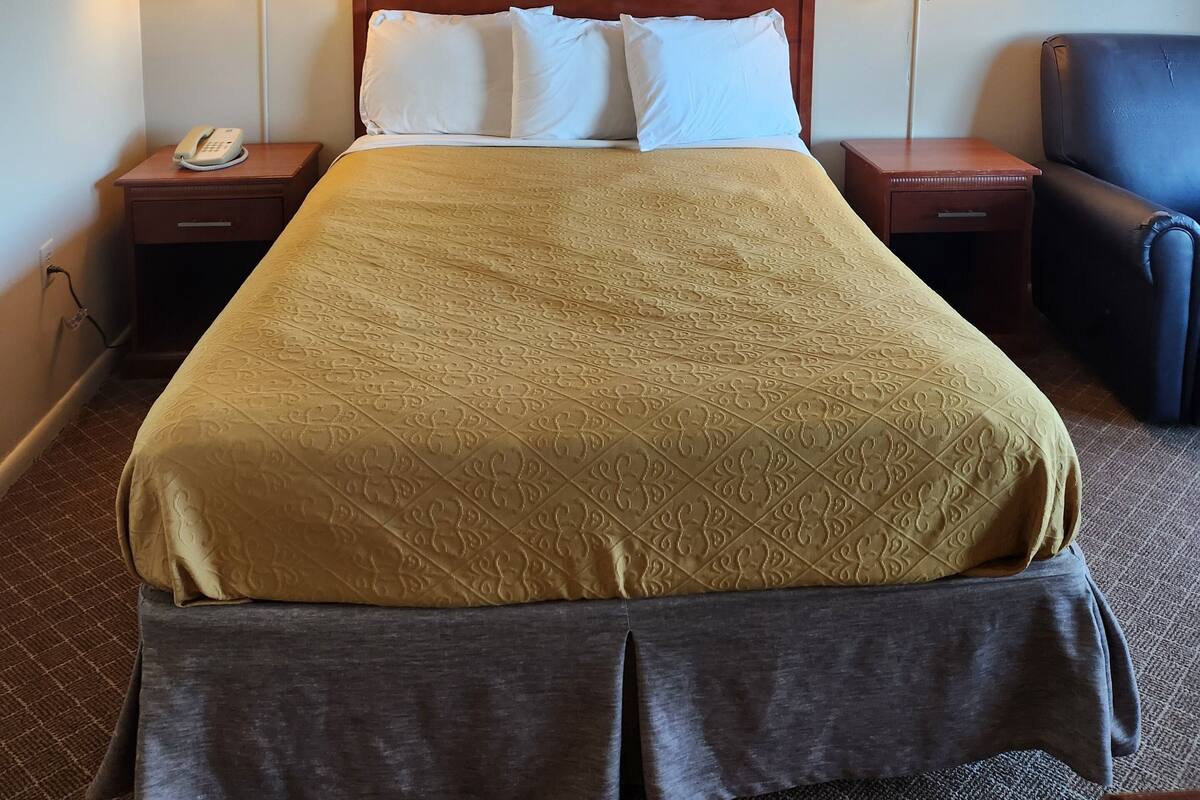 A hotel room with a large bed, two bedside lamps, a telephone, a framed picture on the wall, and a chair.