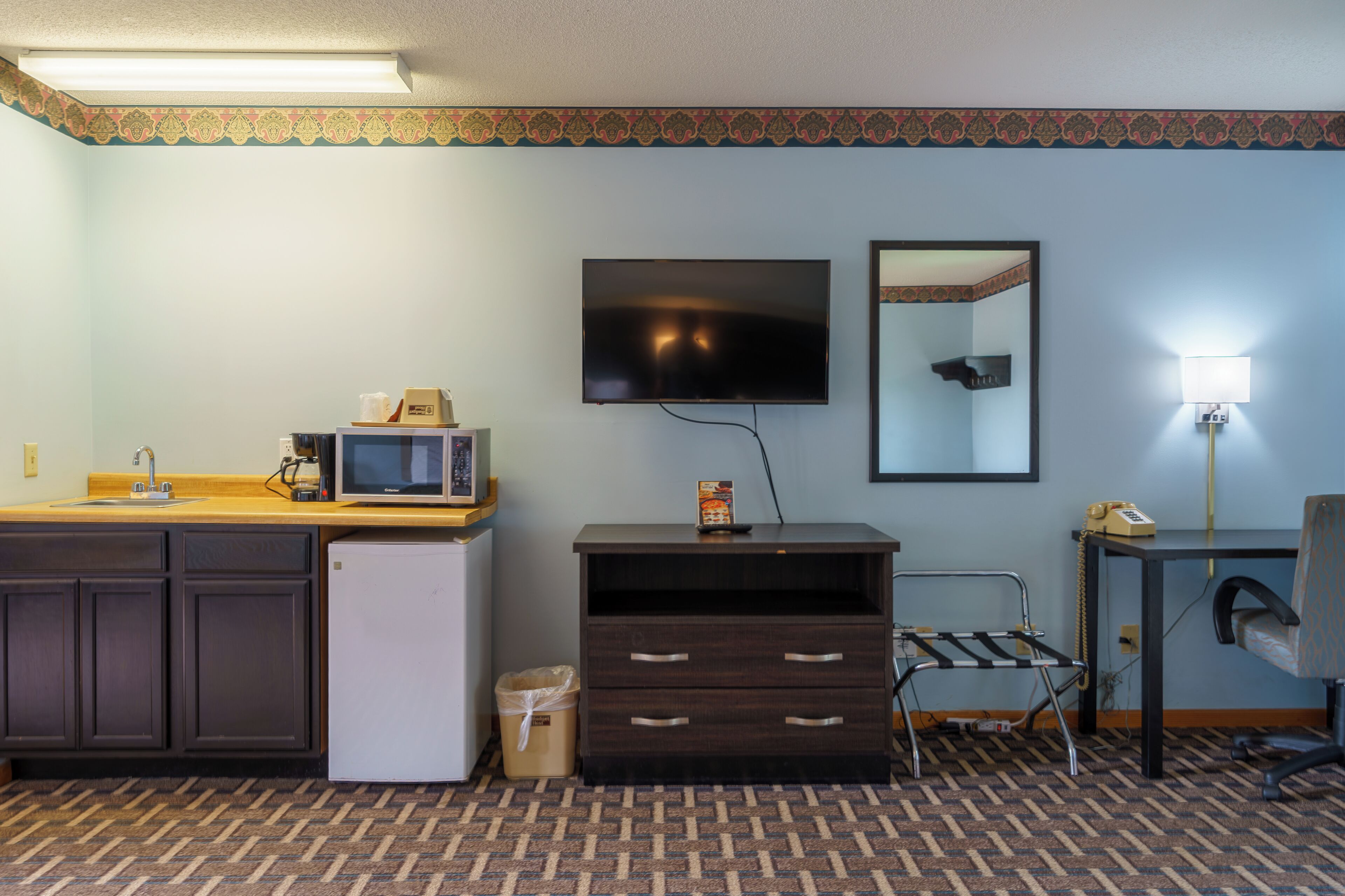 A hotel room with a kitchenette, a flat-screen TV, a mirror, a desk with a lamp, and a patterned carpet.