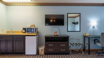 A hotel room with a kitchenette, a flat-screen TV, a mirror, a desk with a lamp, and a patterned carpet.