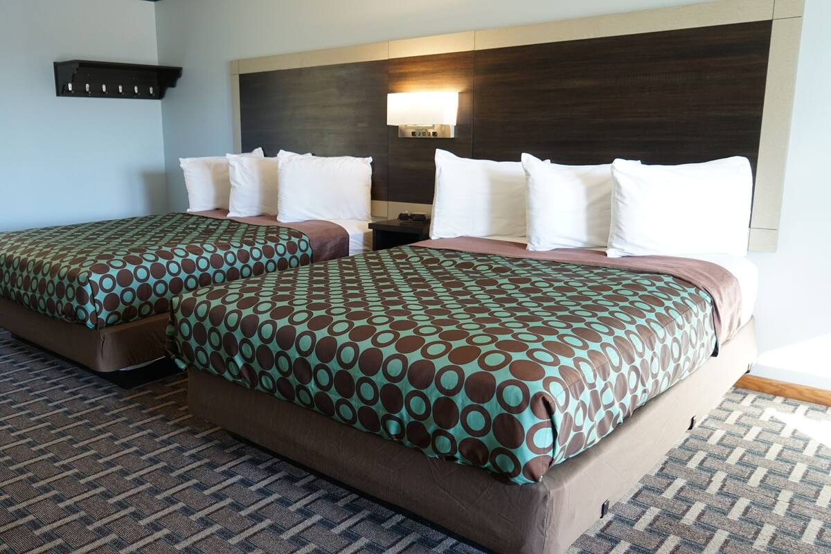 A hotel room with two beds, a large window, a wall-mounted heater, and a nightstand with a lamp.
