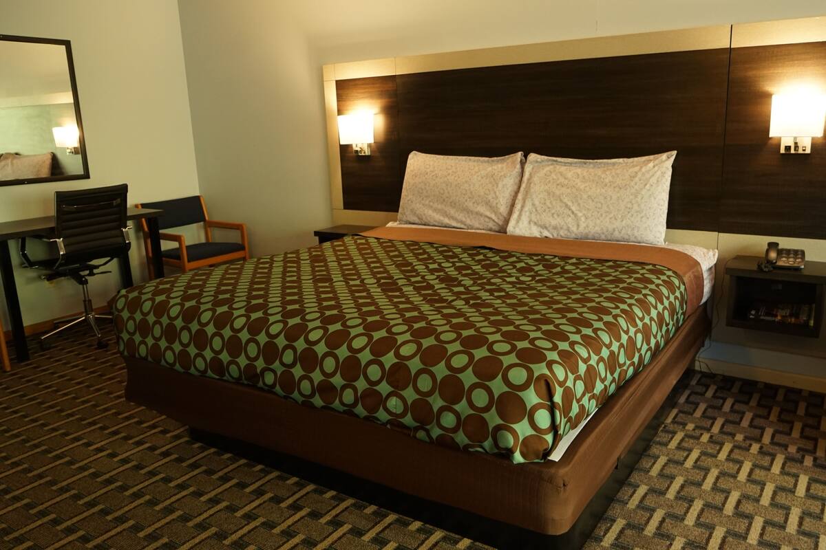 A hotel room with a large bed, a desk, a chair, and a nightstand with a lamp.
