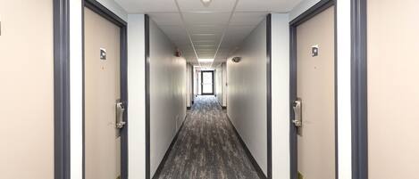 A long, narrow hallway with carpeted floors and doors on both sides, leading to a bright exit at the end.