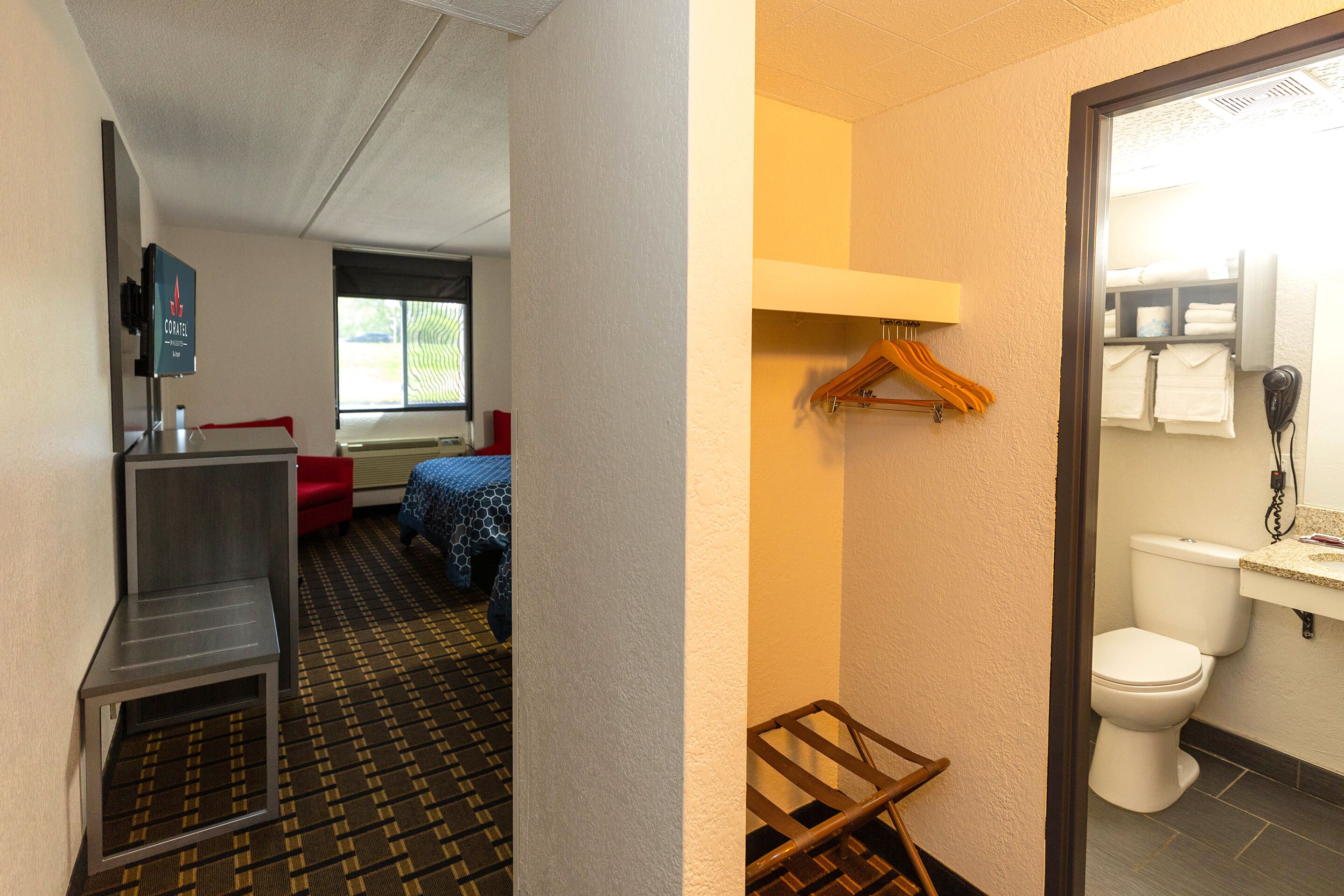 A hotel room with a bed, a small desk, a chair, a hanging coat rack, a bathroom with a toilet, a sink, and a mirror.