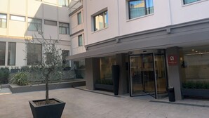 A modern building with a glass entrance, a small tree in a planter, and a sign indicating a business or office space.
