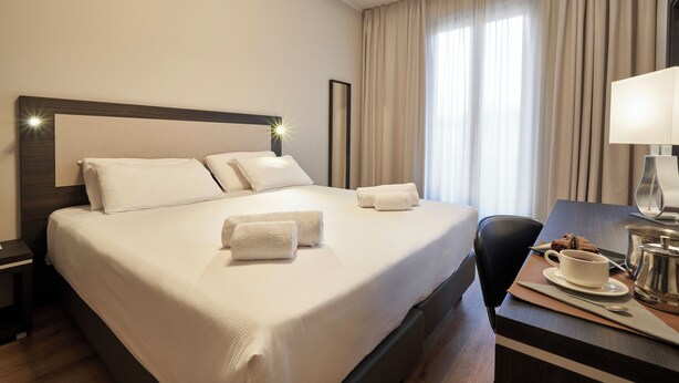 A hotel room with a large bed, a desk with a cup and a pot, and a chair.