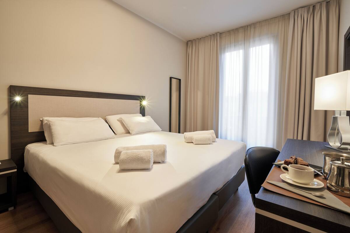 A hotel room with a large bed, a desk with a cup and a pot, and a chair.