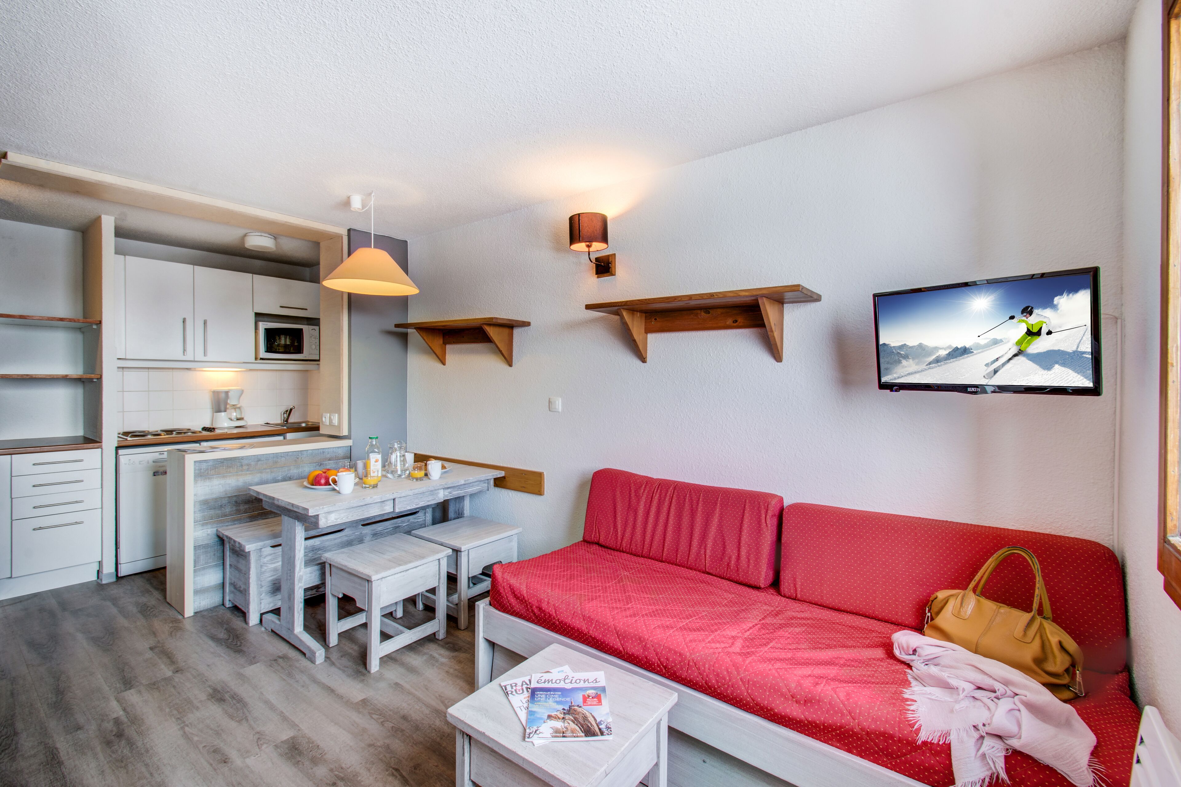 A compact living space with a kitchenette, a dining area, a red sofa, and a TV mounted on the wall.