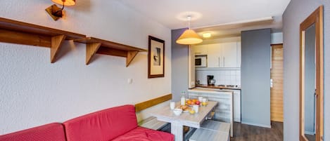 A compact living space with a red sofa, a small dining table with breakfast items, a kitchen area, and a wooden beam shelf.