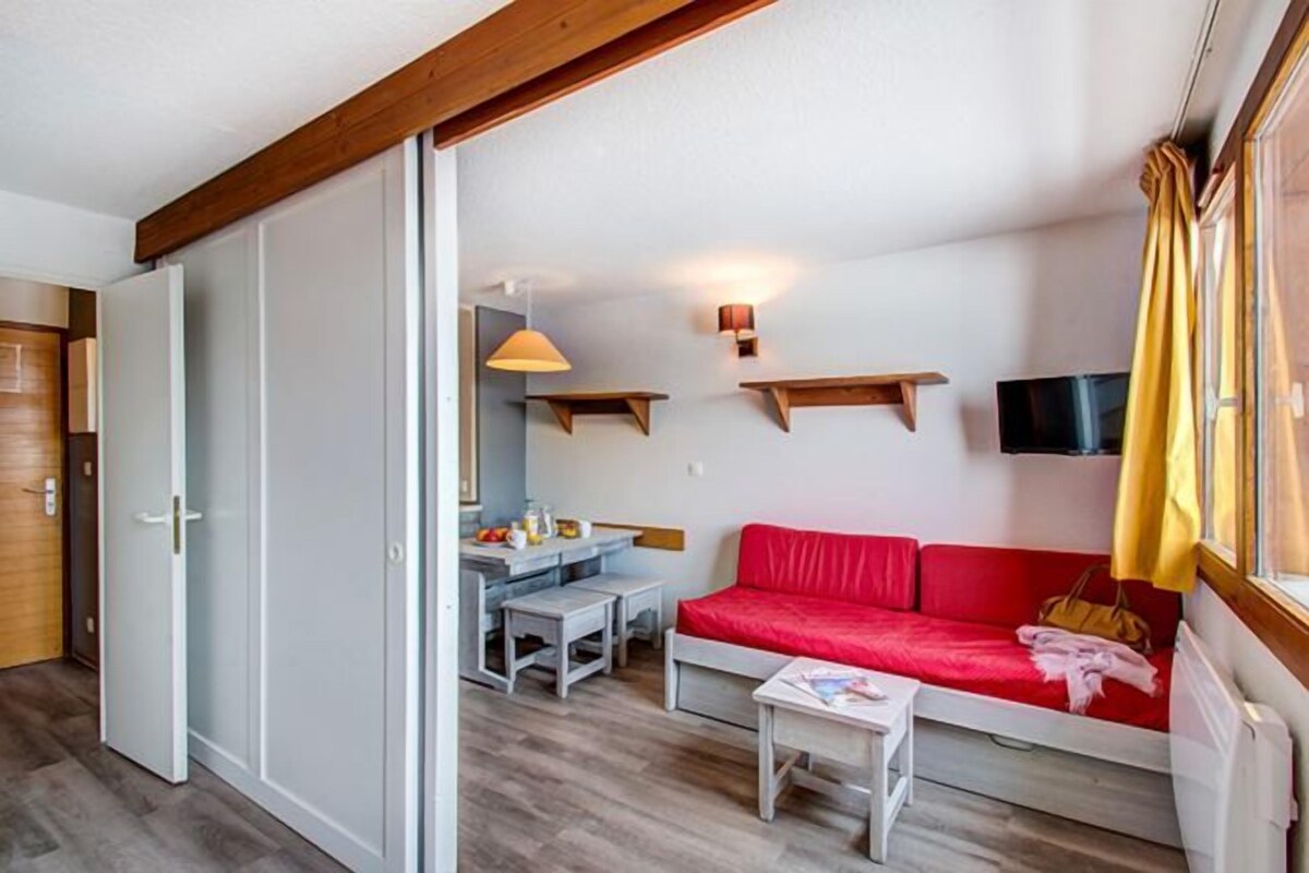 Una habitación compacta con un sofá rojo, una mesita pequeña, un televisor montado en la pared y una viga de madera en el techo.