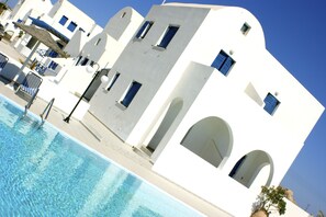 A poolside view of a white building with blue accents, including windows and doorways, and a clear blue sky above. - Hotel Blue Bay Villas (Santorini)