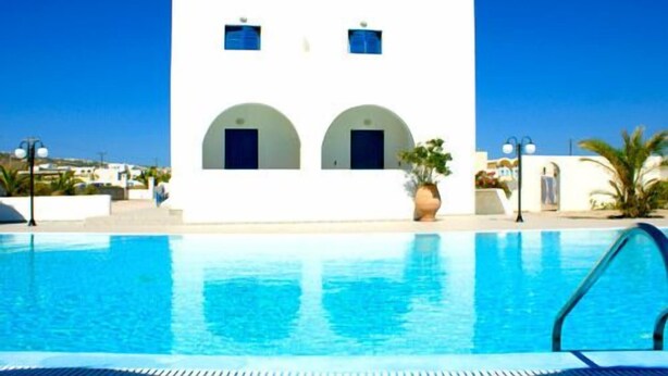 A white building with two arched doorways and a swimming pool in the foreground. - Hotel Blue Bay Villas (Santorini)