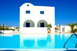 A white building with two arched doorways and a swimming pool in the foreground. - Hotel Blue Bay Villas (Santorini)