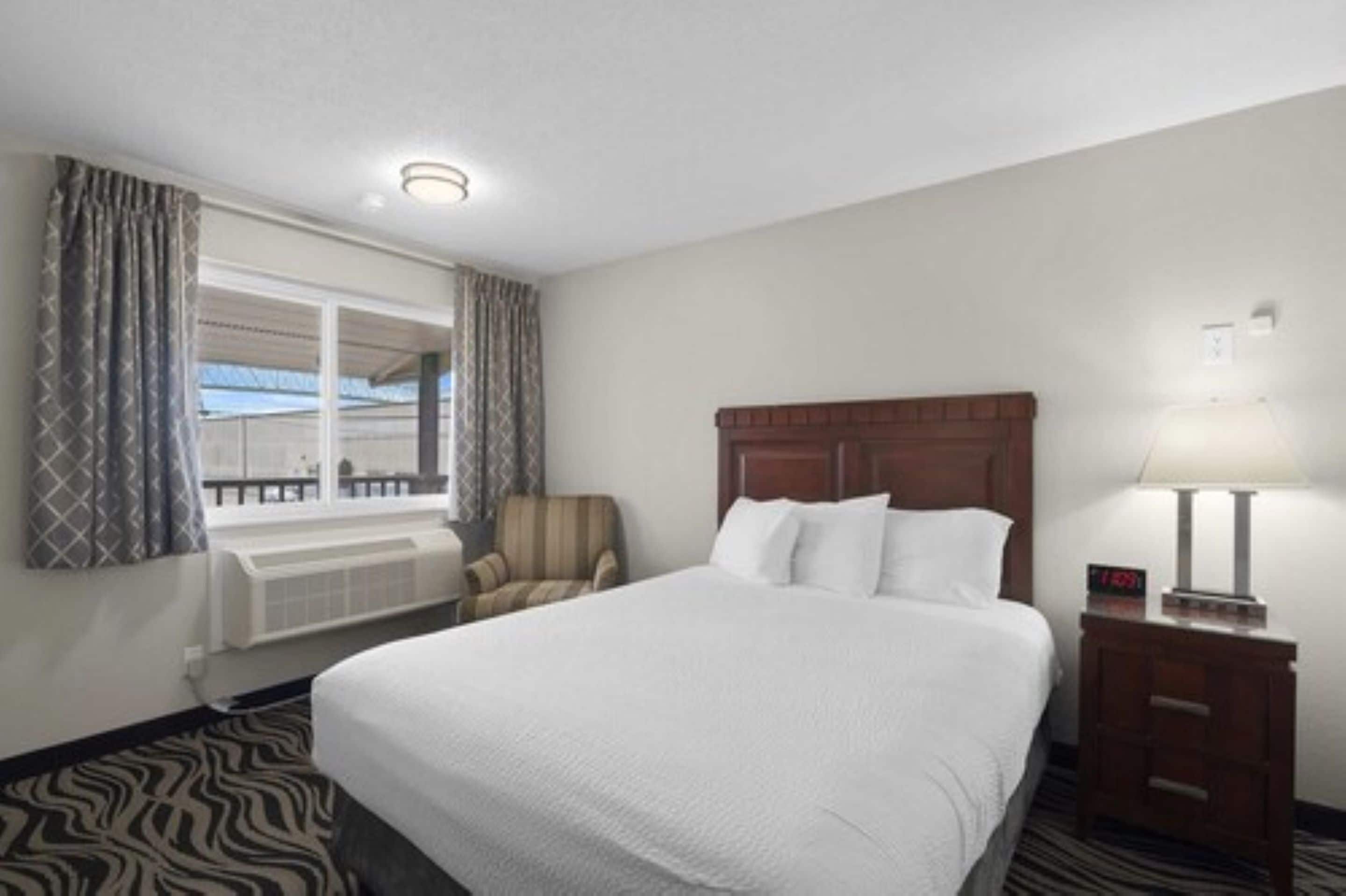 Quarto de hotel com uma cama grande, uma cadeira, uma mesinha de cabeceira com abajur, janela com cortinas e vista para o exterior.