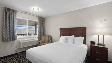 Quarto de hotel com uma cama grande, uma cadeira, uma mesinha de cabeceira com abajur, janela com cortinas e vista para o exterior.