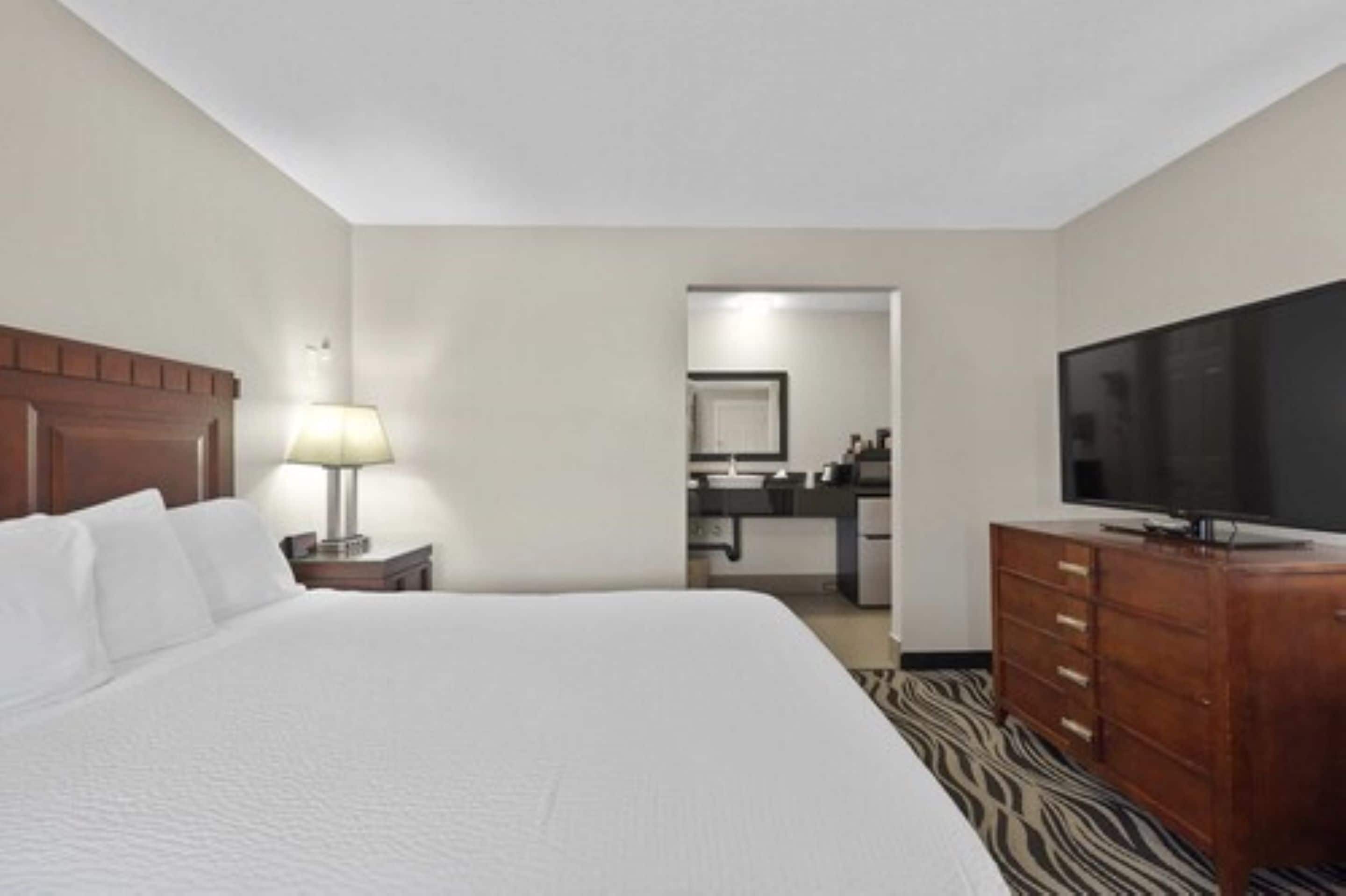 A hotel room with a large bed, a television, a wooden dresser, and a lamp.
