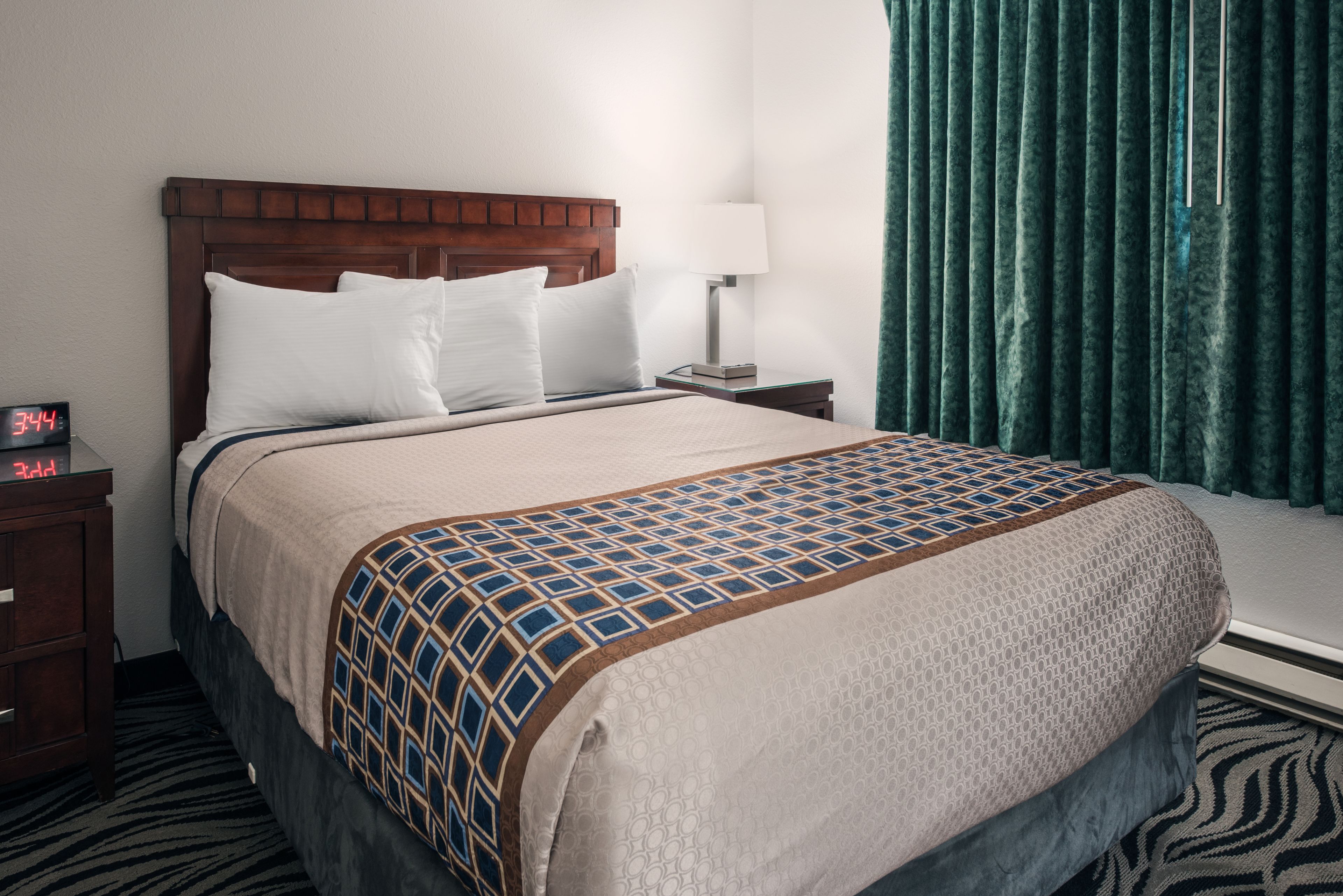 Een hotelkamer met een houten hoofdeinde, een bed met een beddegoed met dessin, een nachtkastje met een lamp en een klok waarop 3:44 staat.
