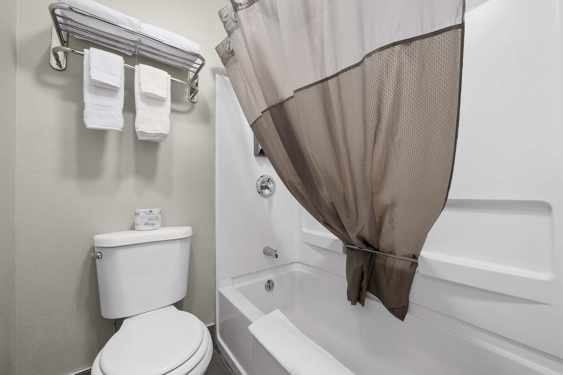 A bathroom with a toilet, a shower with a beige shower curtain, and a towel rack with three white towels.