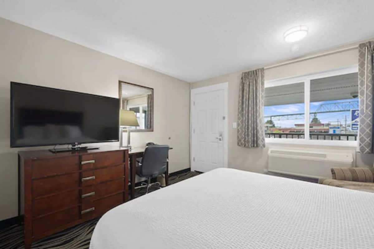 A hotel room with a large bed, a TV mounted on the wall, a desk with a chair, and a window with curtains.