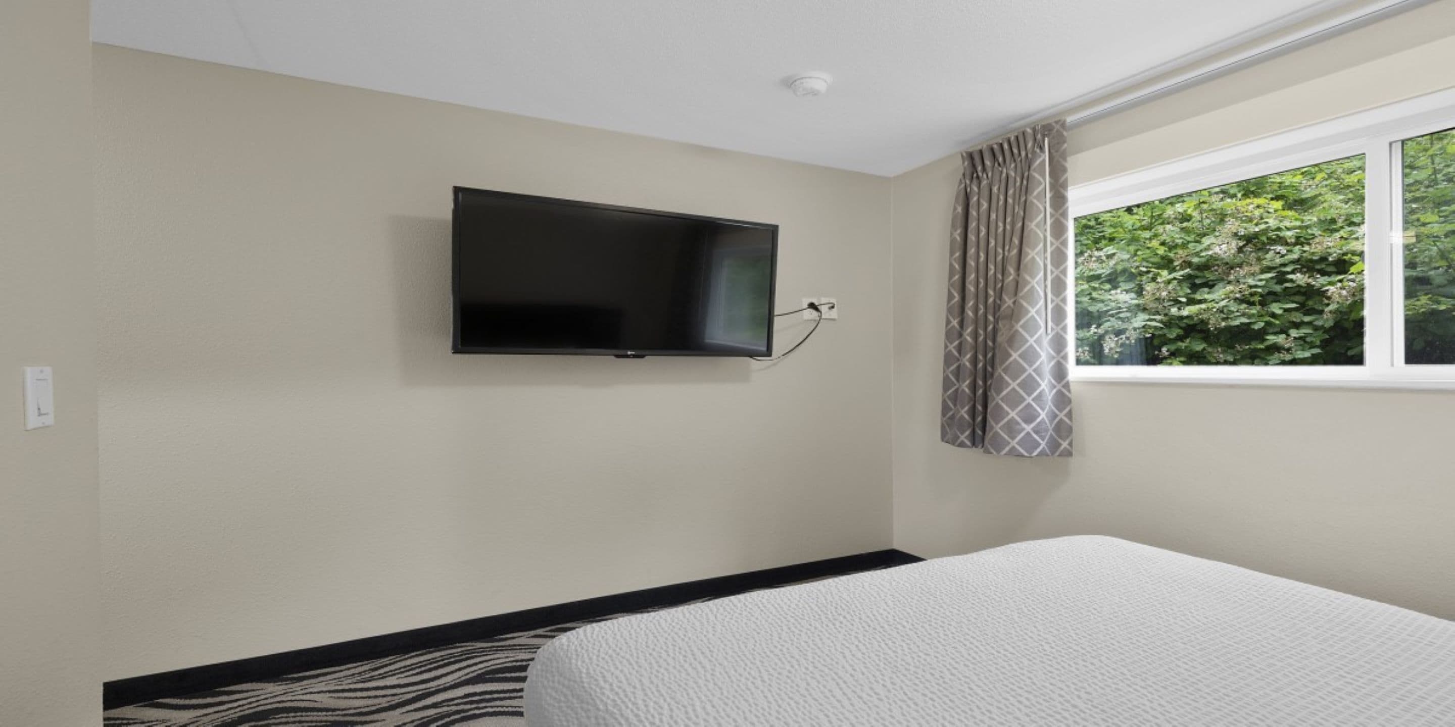 Une chambre d’hôtel avec un lit, un téléviseur fixé au mur et une fenêtre avec des rideaux.