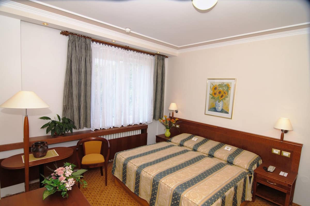 A hotel room with two beds, a desk, a chair, a small table with a vase of flowers, and a window with curtains.