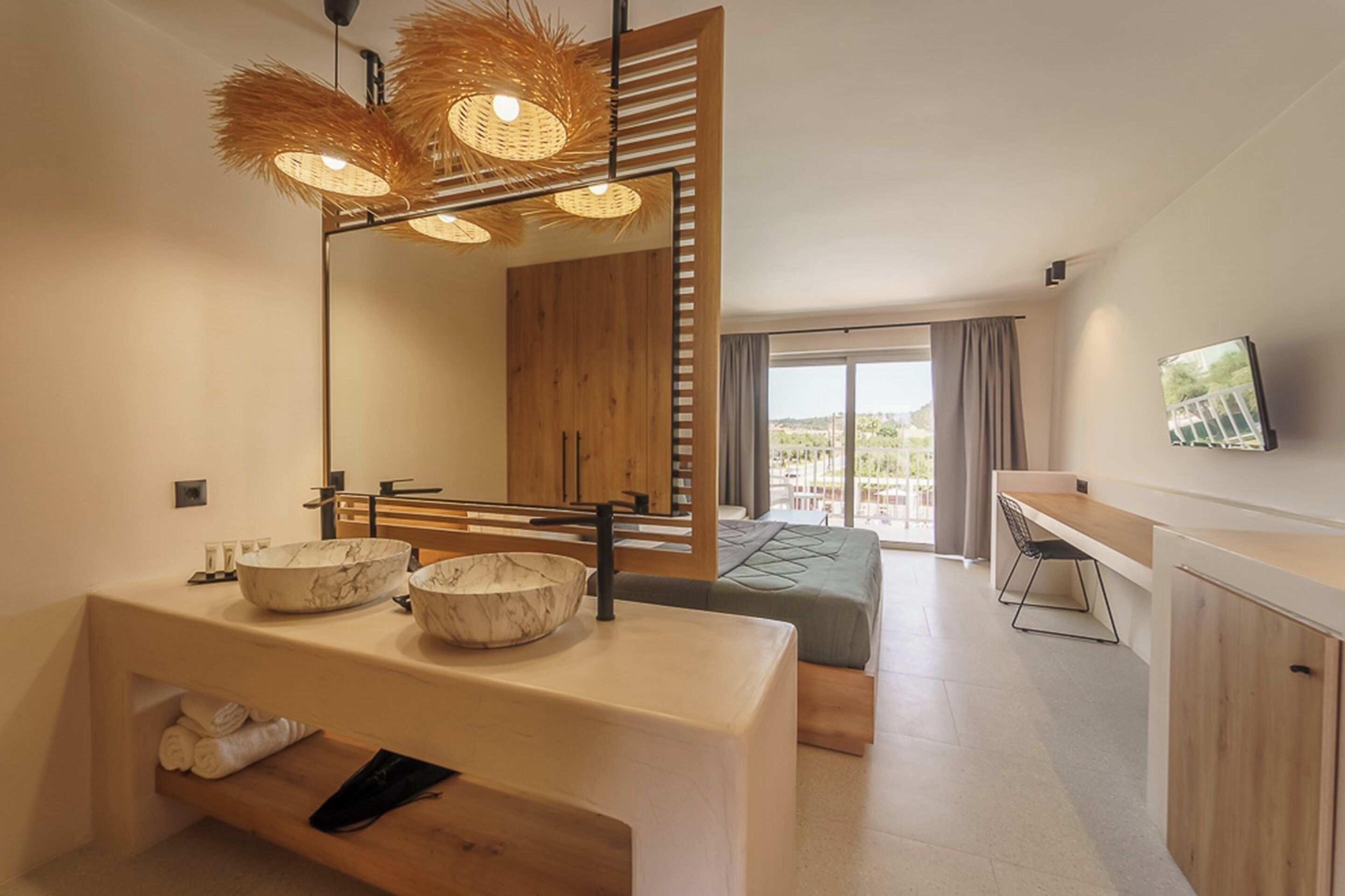 Habitación de hotel moderna con un espejo grande, dos lavamanos de mármol, un tocador de madera, una cama con colcha verde, un escritorio y un ventanal con vistas.