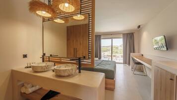 Une chambre d’hôtel moderne dotée d’un grand miroir, de deux vasques en marbre, d’une coiffeuse en bois, d’un lit recouvert d’une courtepointe verte, d’un bureau et d’une grande fenêtre offrant une vue dégagée.
