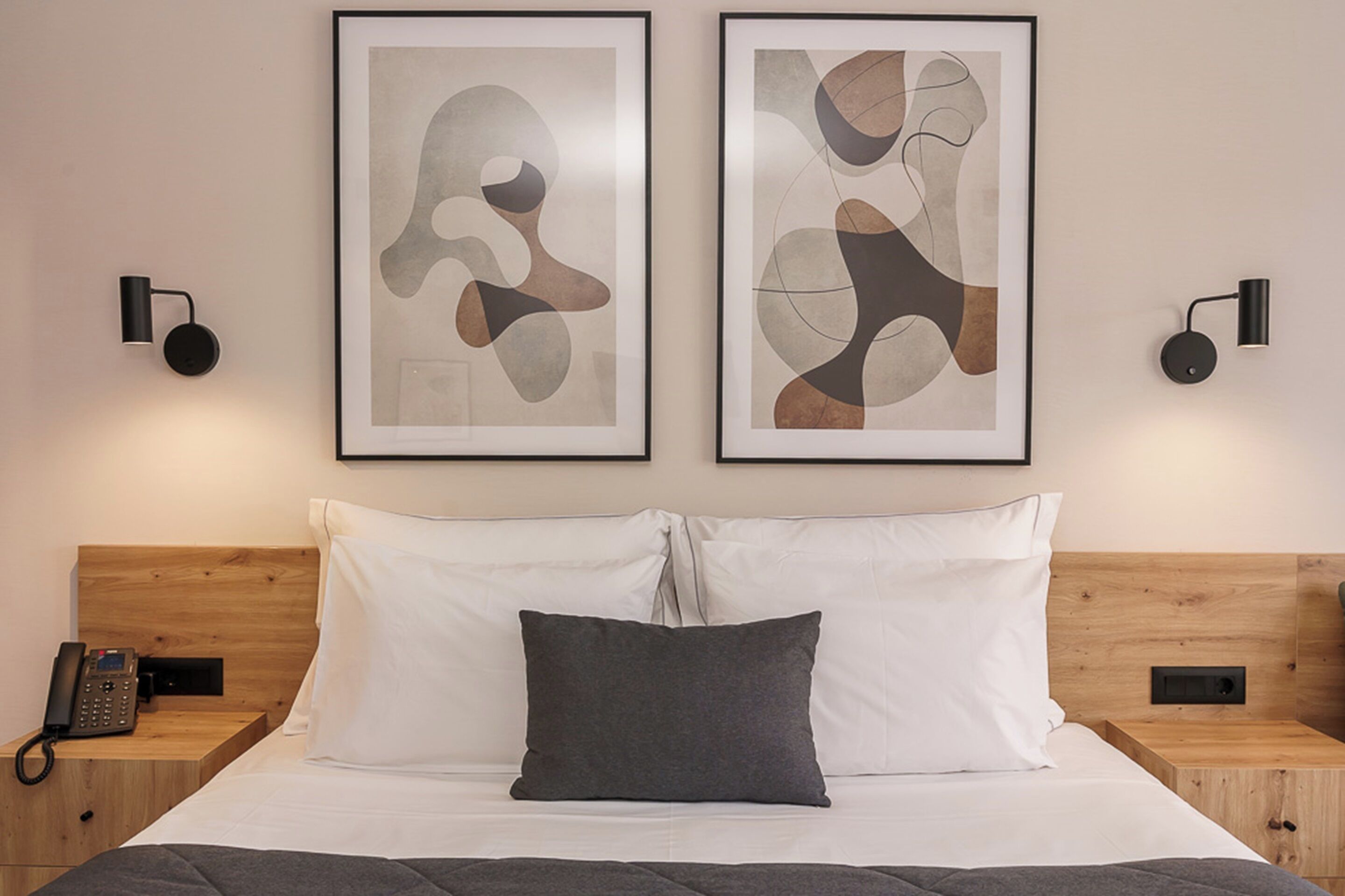 Habitación de hotel con cabecera de madera, cama con ropa de cama blanca y gris, dos obras de arte abstractas enmarcadas, un teléfono en la mesita de noche y dos lámparas fijadas a la pared.