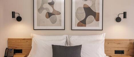 Habitación de hotel con cabecera de madera, cama con ropa de cama blanca y gris, dos obras de arte abstractas enmarcadas, un teléfono en la mesita de noche y dos lámparas fijadas a la pared.