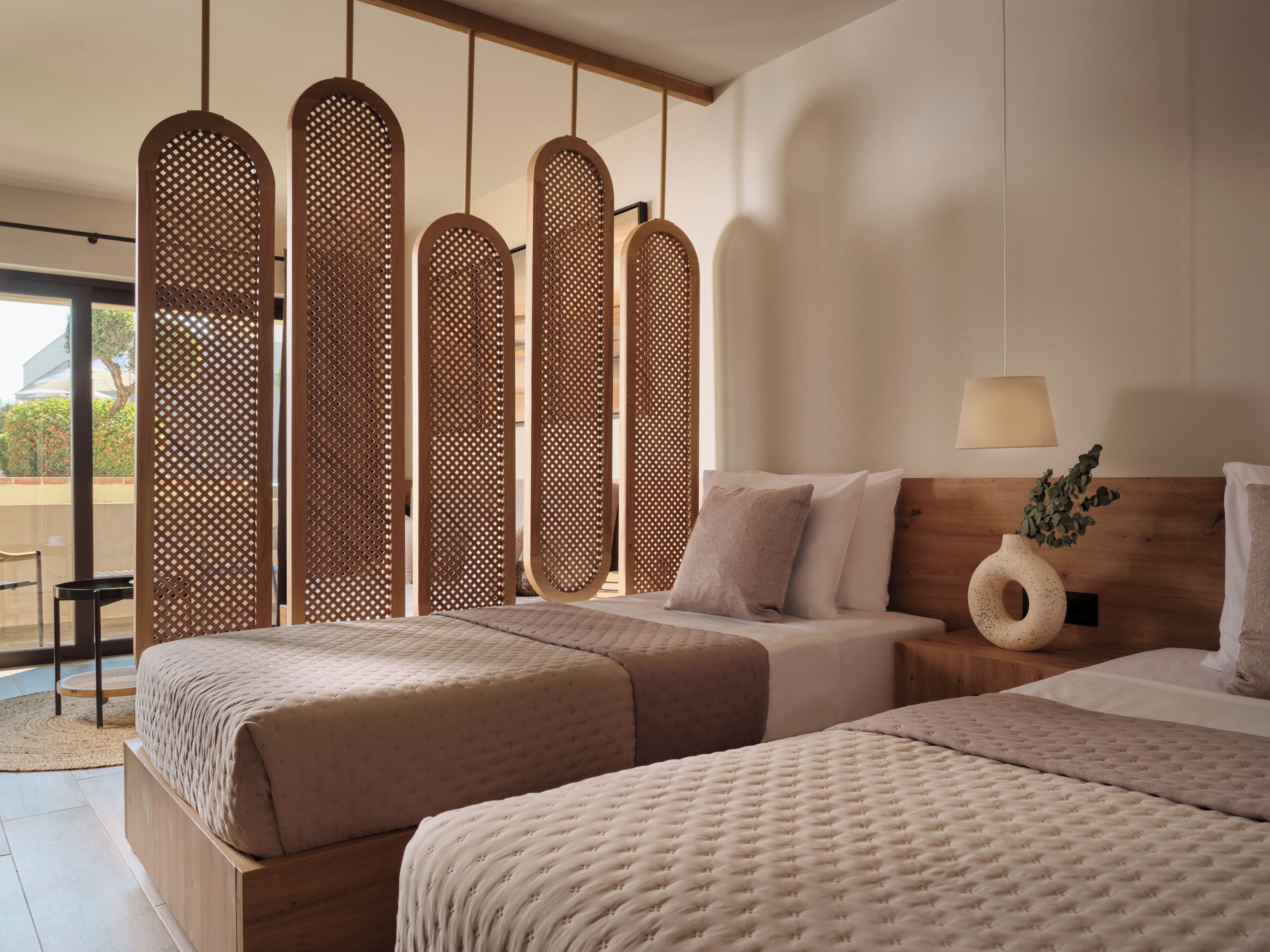 Habitación de hotel con dos camas, cabecera de madera, mesita de noche con lámpara y un florero decorativo.