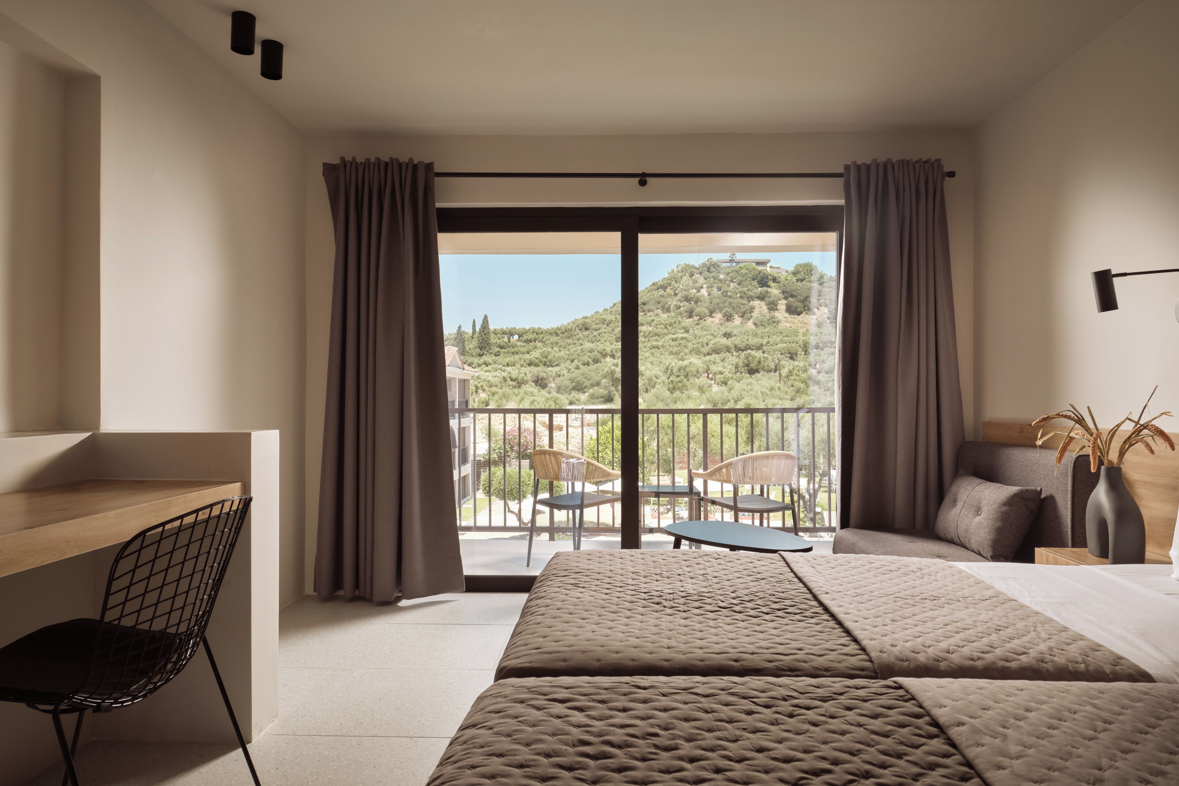 Une chambre d’hôtel avec un lit, un bureau et une chaise. Elle dispose d’un balcon offrant une vue sur la verdure et les bâtiments.