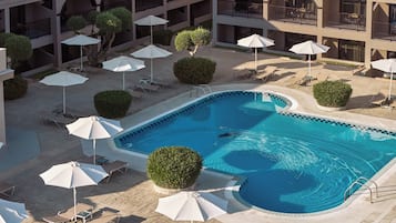 Ein Swimmingpool, umgeben von Liegestühlen und Sonnenschirmen, mit einem mehrstöckigen Gebäude im Hintergrund.