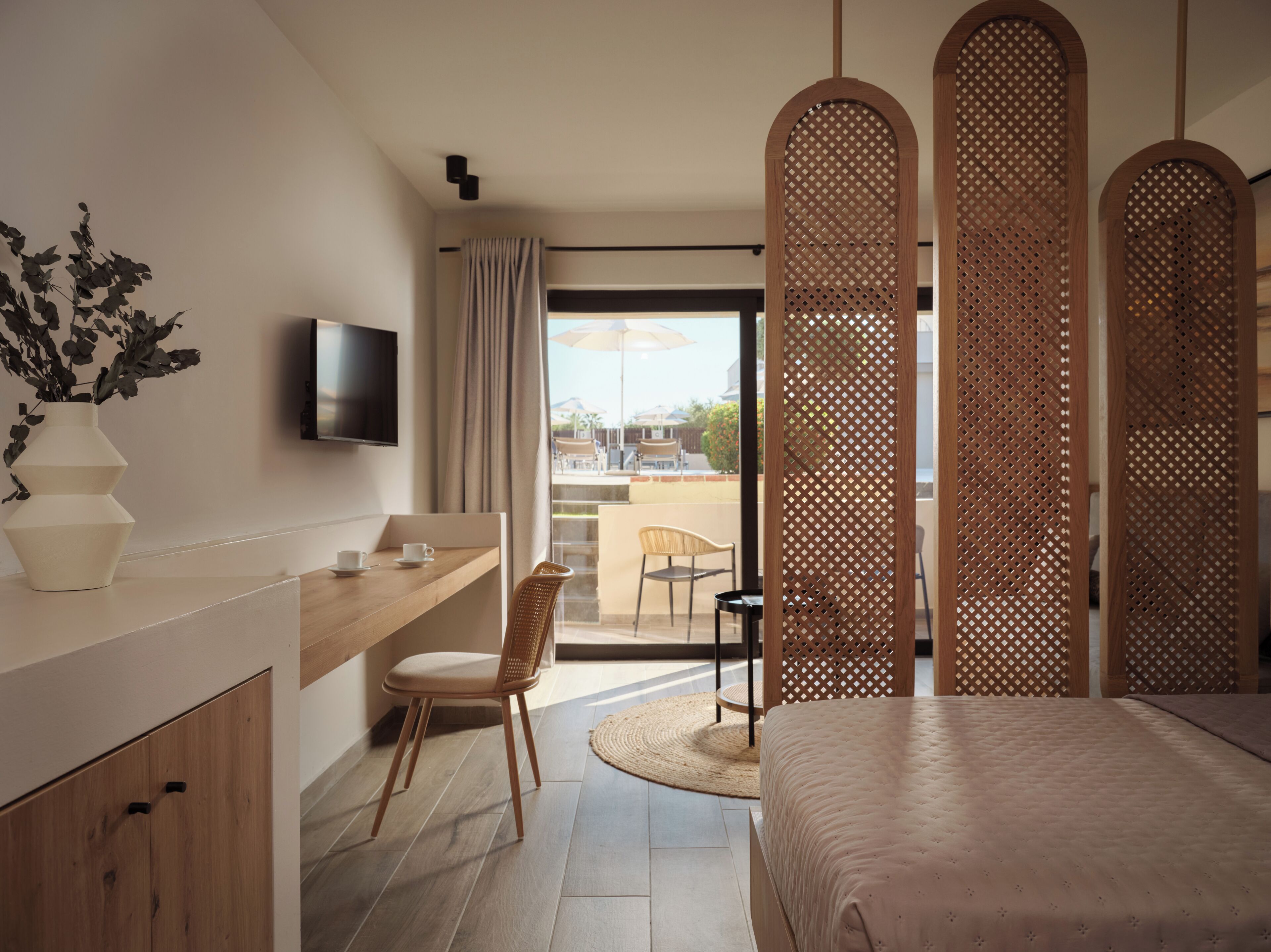A modern hotel room with a wooden desk, a bed, a TV, and a balcony with outdoor seating.