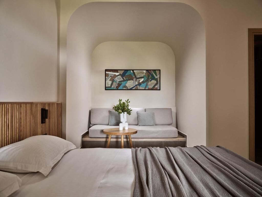 Una habitación de hotel moderna con cama, sofá, una mesita con un florero y un cuadro en la pared.