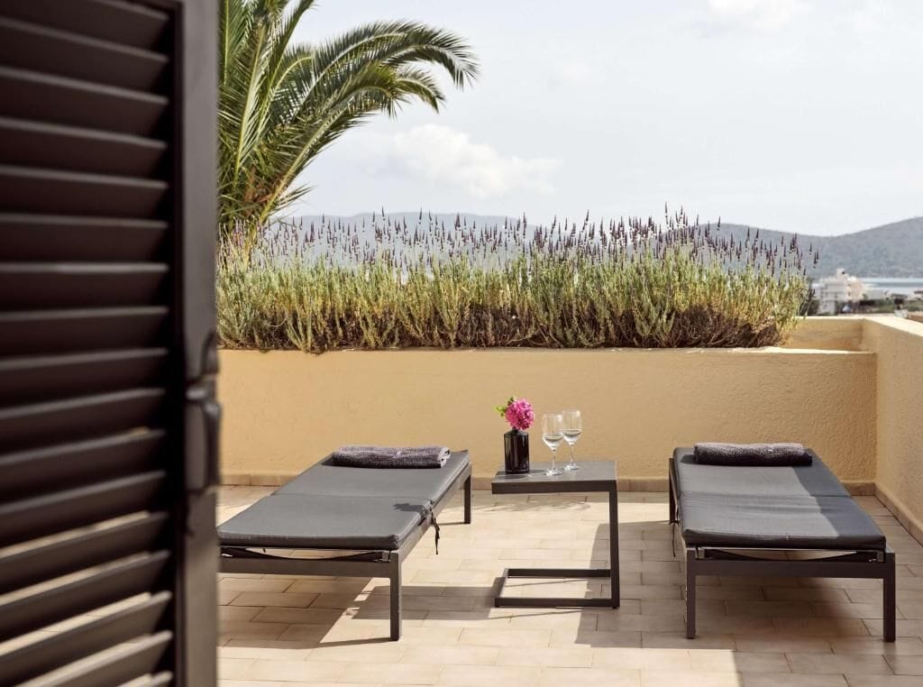 Une terrasse avec deux chaises longues, une table ornée d’un vase de fleurs et de deux verres, et une vue sur la mer et les collines.