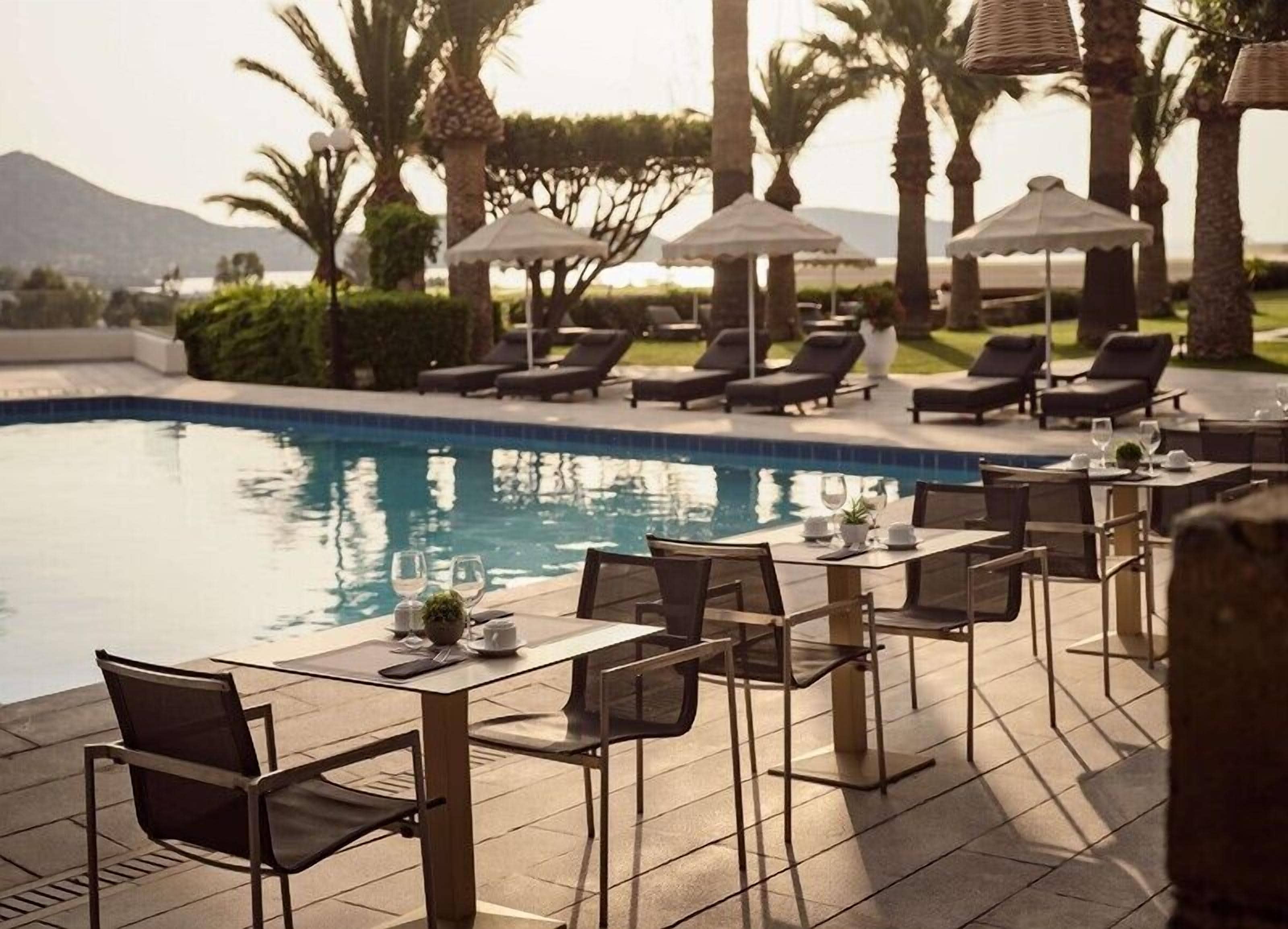 A poolside dining area with tables and chairs, umbrellas, and lounge chairs.