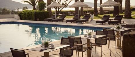 Un área de comedor junto a la piscina con mesas, sillas, sombrillas y sillones de descanso.