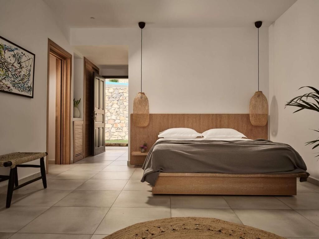 A modern bedroom with a wooden headboard, a bench, a bed with a grey bedspread, and two hanging pendant lights.