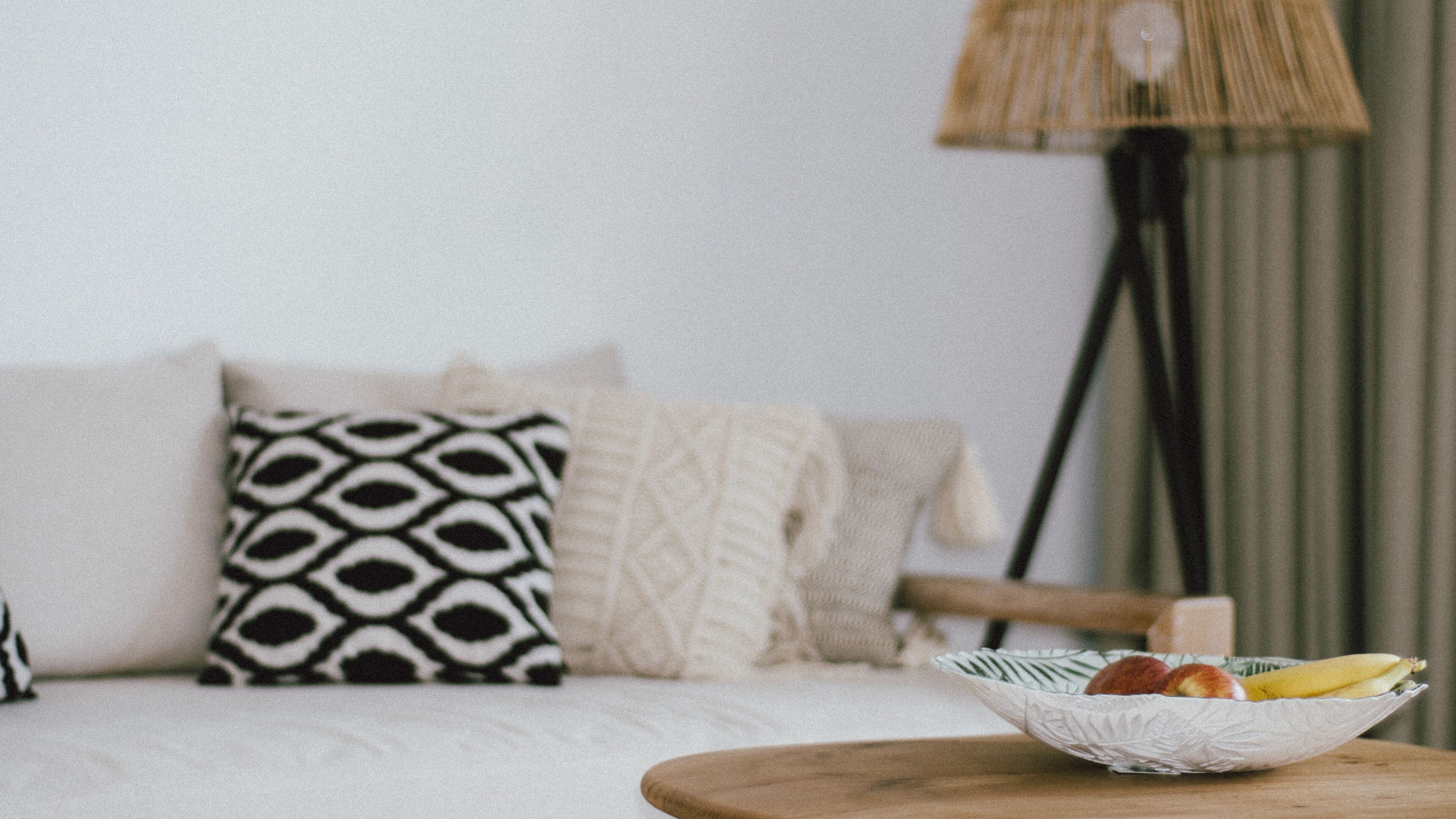A cozy living room with a sofa, patterned pillows, a lamp, and a bowl of fruit on a side table.