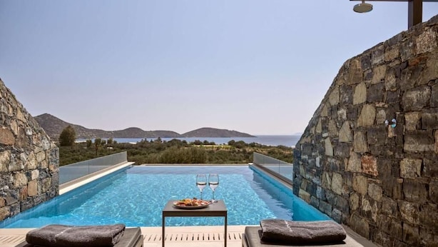 A private pool area with a stone wall, lounge chairs, and a table with a glass of wine and a plate of food. - Elounda Palm Hotel (Agios Nikolaos)