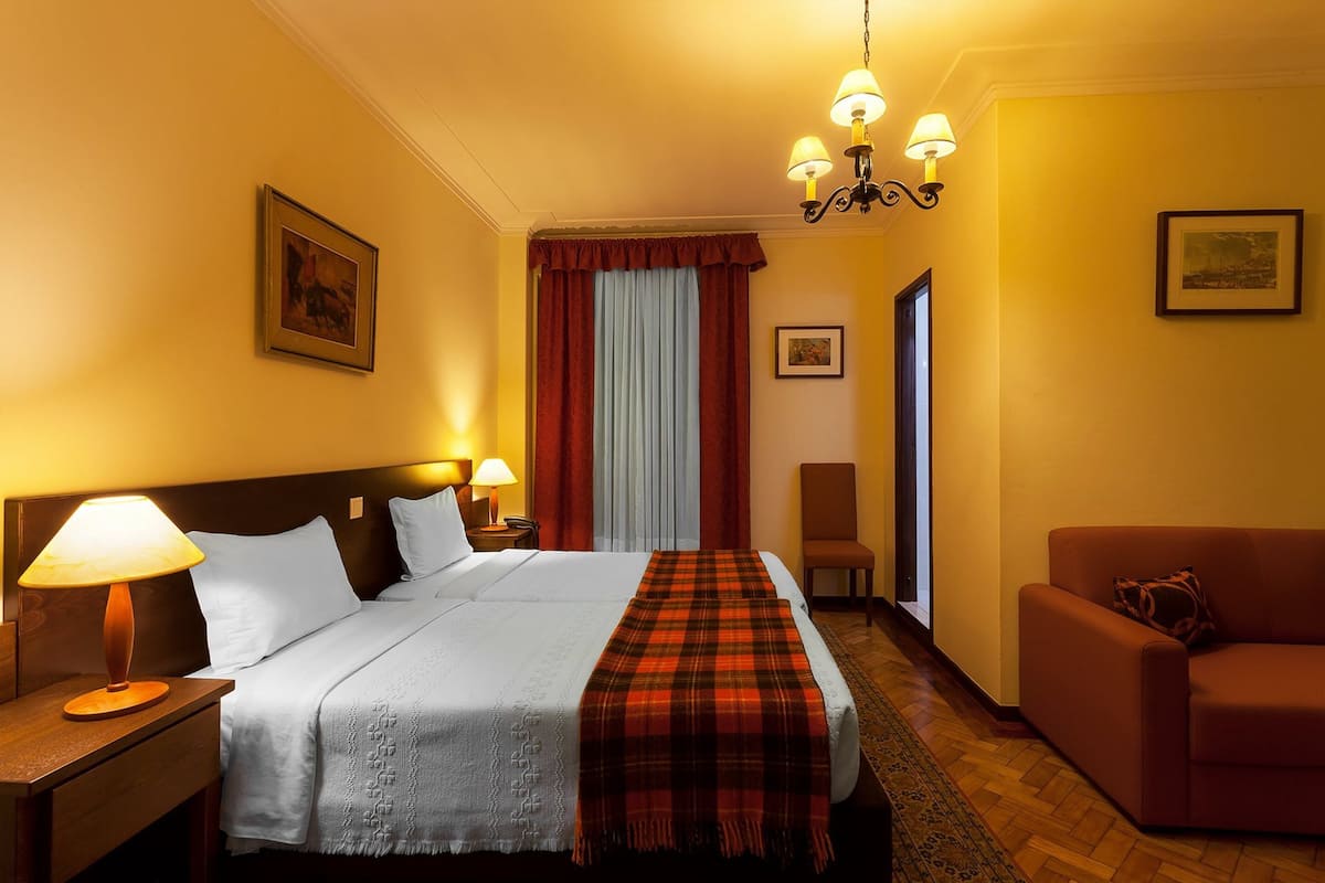 A hotel room with a large bed, two bedside lamps, a red plaid throw blanket, a wooden nightstand, a brown armchair, and a chandelier.